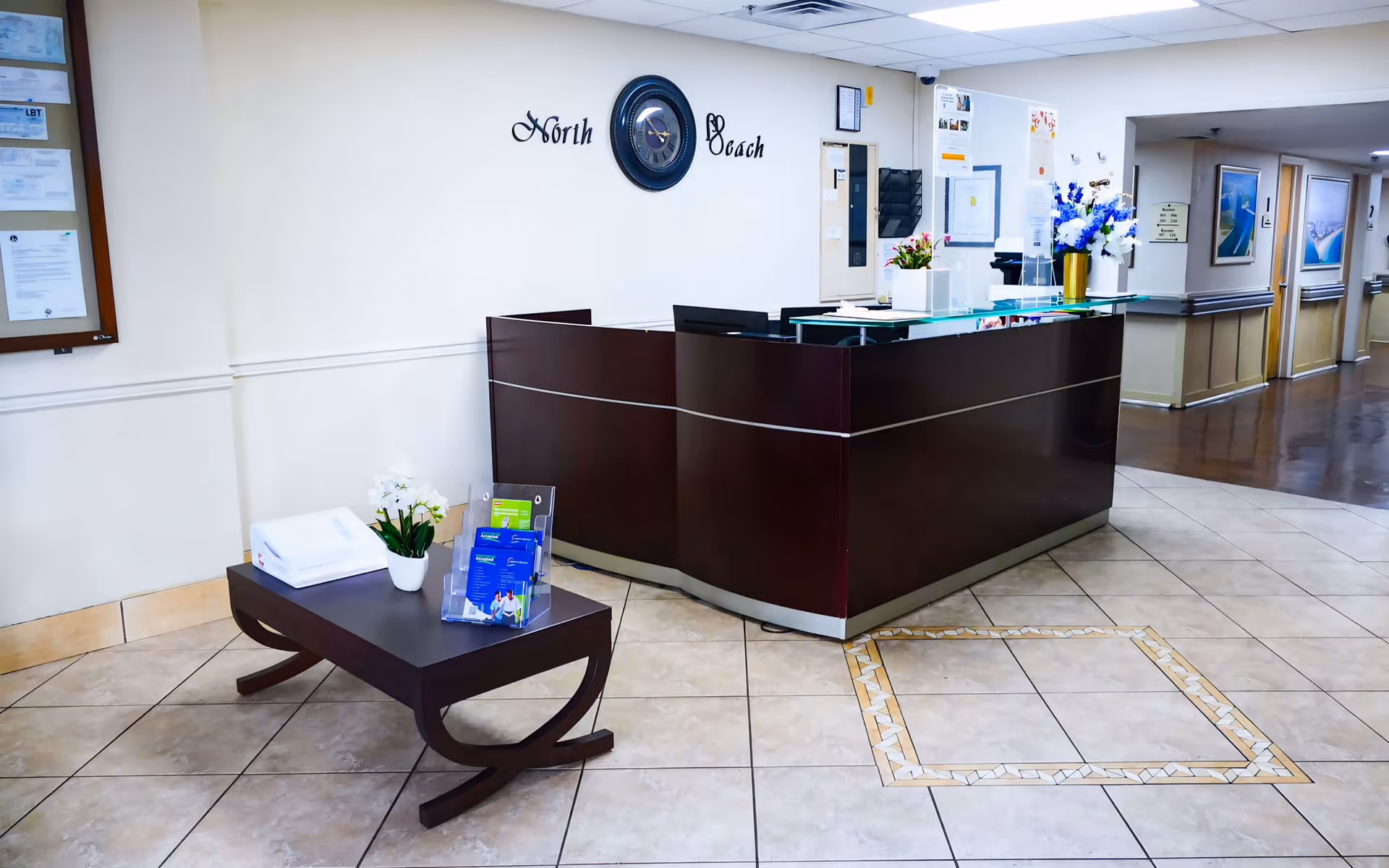 Reception area of North Beach Rehabilitation Center with a dark wood reception desk, a wall clock above with the words 'North Beach', a small table with brochures and a potted plant, and a hallway leading to other rooms.