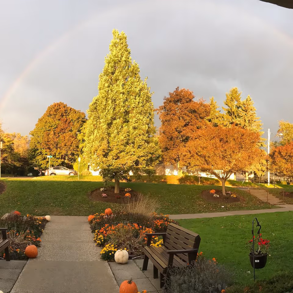 Pathway and benches in a landscaped lawn with pumpkins, fall trees, and a faint rainbow overhead.