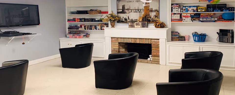 A cozy common area with five black armchairs arranged in a semi-circle facing a wall-mounted flat screen TV. The room features a white fireplace with a brick interior, flanked by built-in white shelves and cabinets filled with books, games, and decorative items. The walls are painted light gray and the floor is light-colored tile.