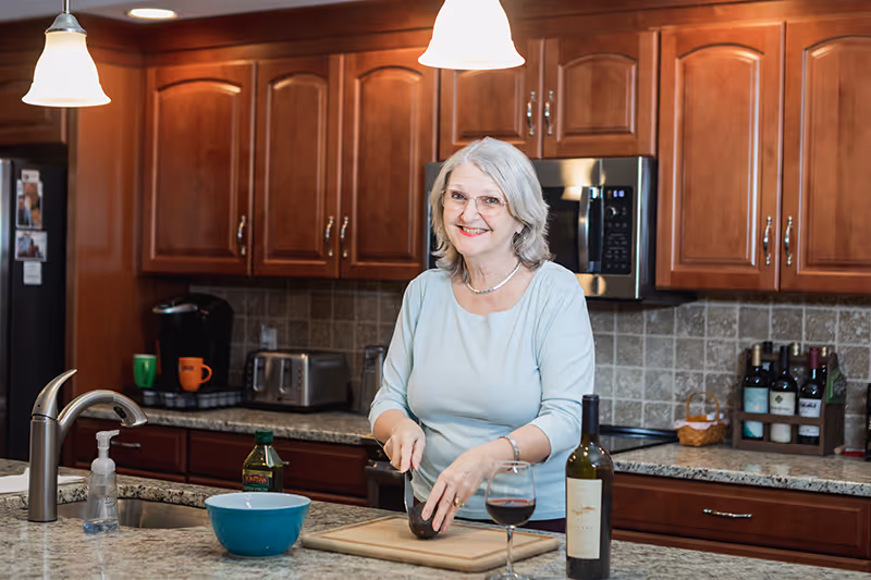 A smiling woman stands at a kitchen island slicing an avocado with a wine glass and bottle nearby.