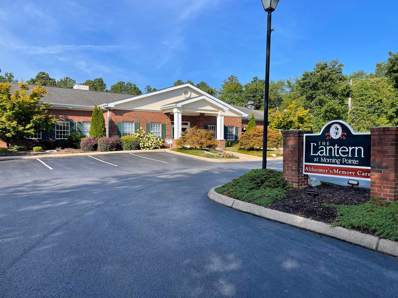 Exterior view of The Lantern at Morning Pointe Alzheimer's Center of Excellence in Collegedale, showing a brick building with white columns at the entrance, surrounded by landscaped bushes and trees under a clear blue sky. A sign in front reads 'The Lantern at Morning Pointe Alzheimer's/Memory Care.'