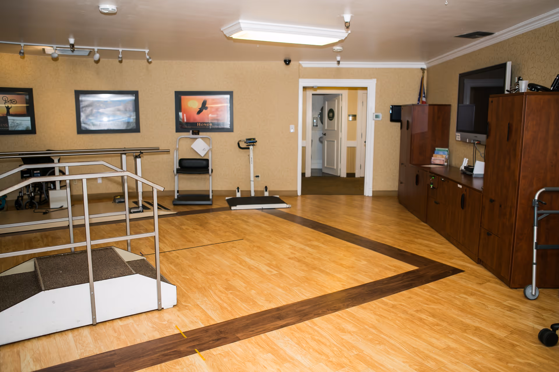An indoor rehabilitation room with wooden flooring and a small ramp with handrails for physical therapy. The room has exercise equipment including a treadmill and a balance scale. There are framed pictures on the wall and wooden cabinets with a television mounted above them. A walker is partially visible on the right side.