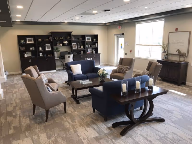 A bright and spacious senior living common area with two blue sofas, four beige armchairs, a dark wooden coffee table with a flower arrangement, and a side table with four candles. In the background, there is a large dark wooden bookshelf with various decorative items and a computer, a window letting in natural light, and a door leading outside.