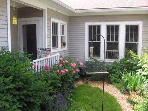 Front of a gray house with a garden path, pink roses, and lush greenery.