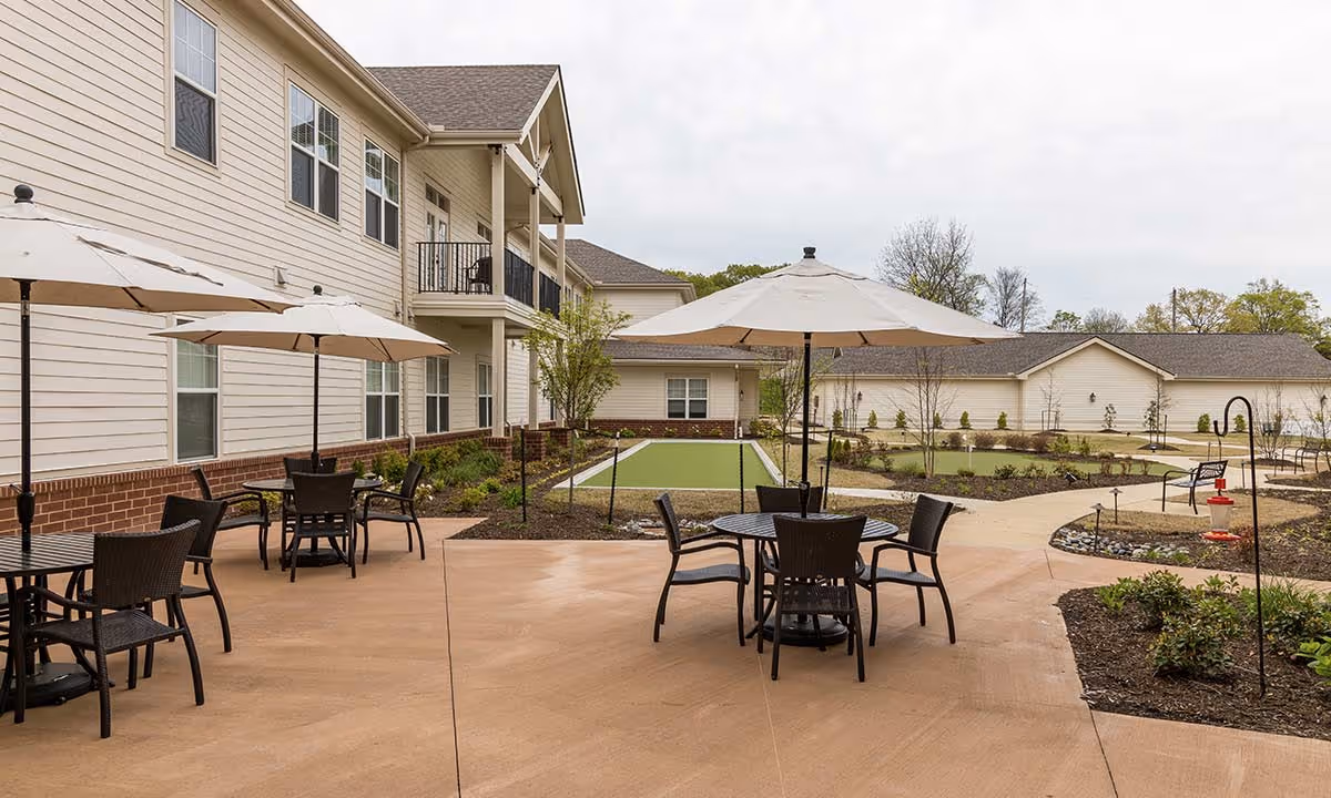 Outdoor patio area at StoryPoint Cordova with round tables and chairs under large beige umbrellas. The patio is adjacent to a two-story building with beige siding and multiple windows. In the background, there are landscaped garden beds, walking paths, and a bocce ball court.