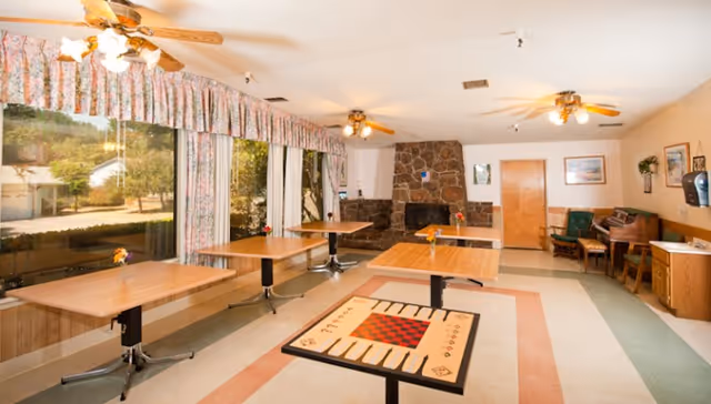 A spacious common room with several wooden tables, one featuring a checkerboard and backgammon design. The room has large windows with floral curtains, ceiling fans with lights, a stone fireplace, and various chairs and furniture along the walls.