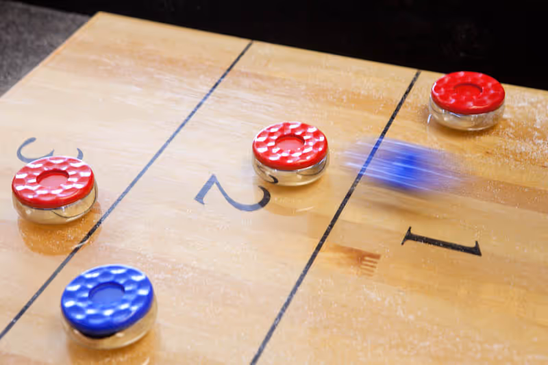 Close-up of a shuffleboard table with red and blue pucks, one blue puck is in motion sliding across the table.