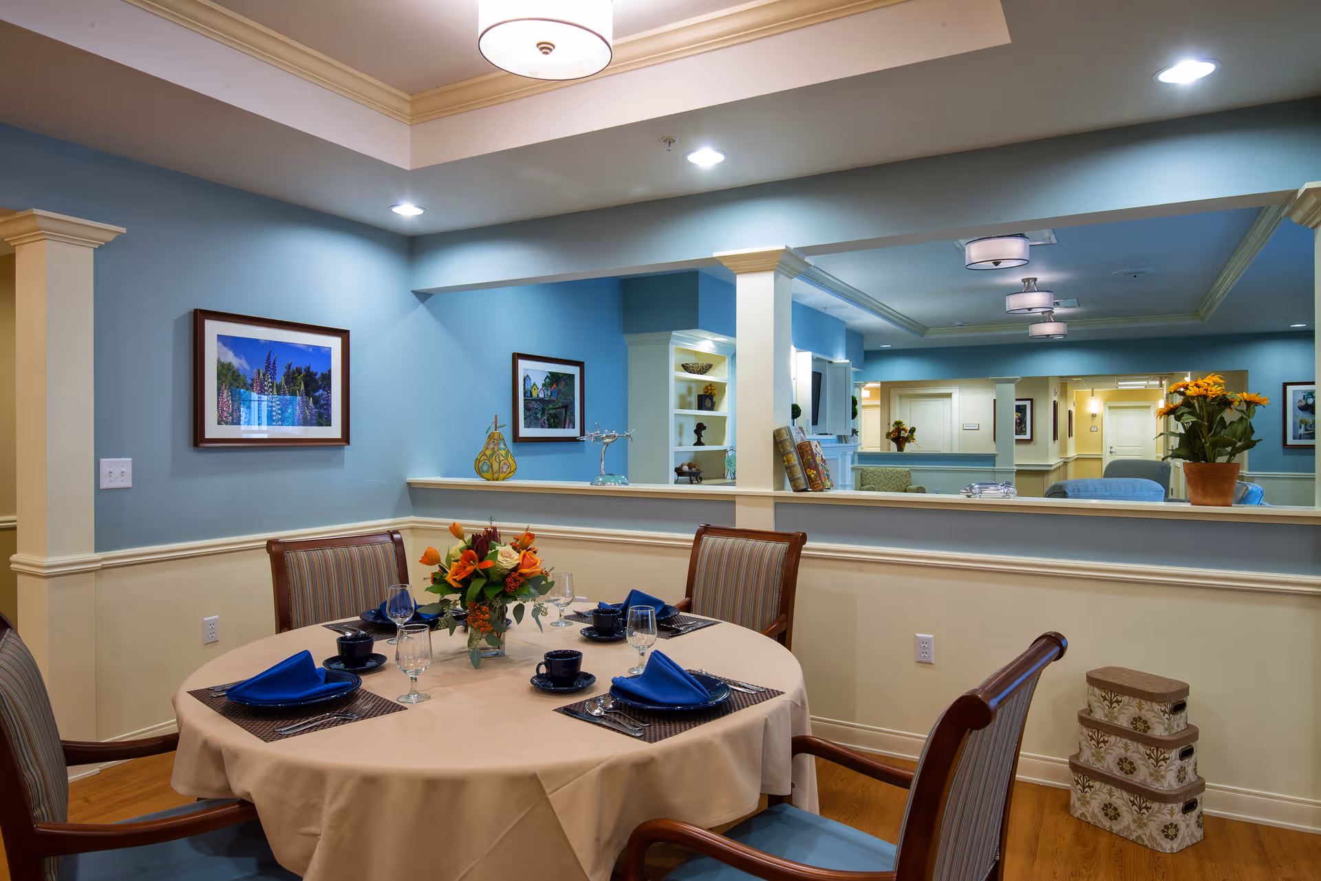 A dining room with a round table covered with a beige tablecloth set for four people with blue napkins, black cups, and clear glasses. The room has light blue walls with cream-colored wainscoting, framed pictures on the walls, and a large mirror reflecting the opposite side of the room. There are wooden chairs with striped cushions around the table, a floral centerpiece, and decorative boxes stacked in the corner.