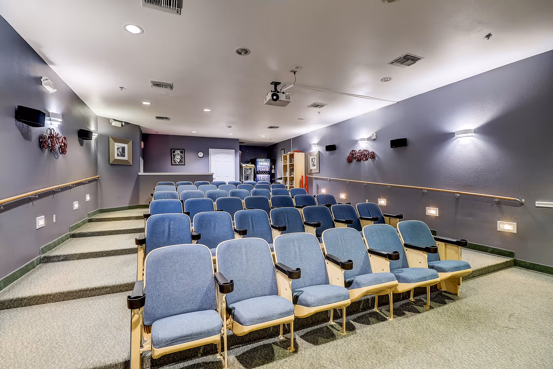 A small theater room with multiple rows of blue cushioned seats facing a projector mounted on the ceiling. The room has gray walls with handrails along the sides, wall-mounted lights, and framed pictures. There is a vending machine and a bookshelf at the back of the room.