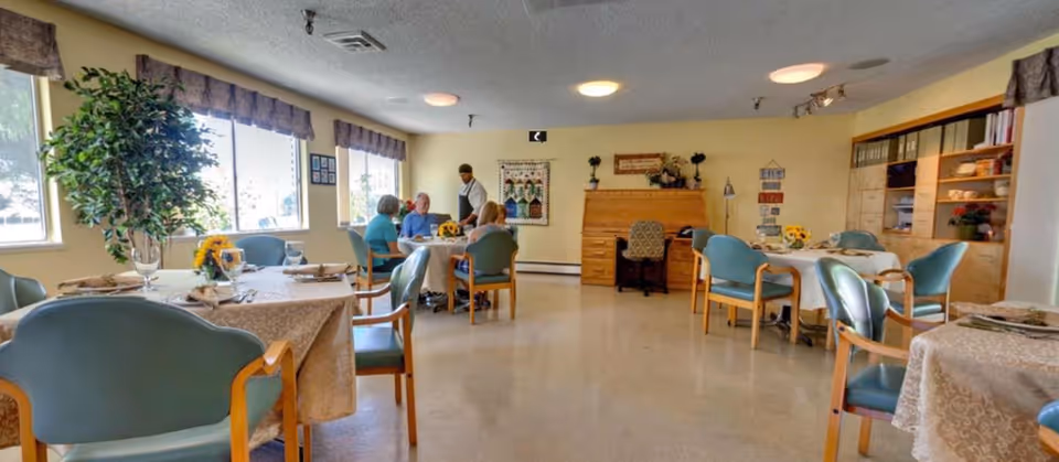 Dining room with tables set for meals, teal chairs, large windows, a potted plant, and a small group of residents talking near the back.