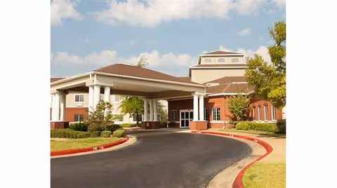 Exterior view of a single-story brick building with a covered entrance driveway, surrounded by landscaped greenery and trees under a partly cloudy sky.