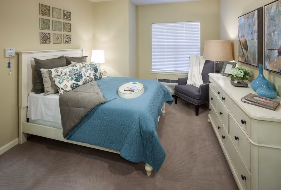 A cozy bedroom with a white wooden bed frame, a blue quilted bedspread, and multiple decorative pillows. There is a white tray with a book and glasses on the bed. A white dresser with black knobs is on the right side, decorated with two blue vases, a small plant, and framed pictures. A gray armchair with a white throw blanket is positioned near a window with closed blinds. The walls are light beige with framed artwork above the bed and on the wall near the dresser. A table lamp is on the dresser, providing warm lighting.