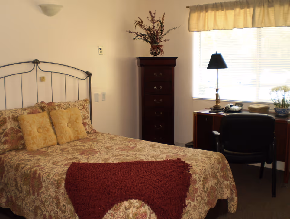 A bedroom with a bed featuring a patterned bedspread and two yellow pillows, a dark wooden tall dresser with a vase of flowers on top, a desk with a lamp, telephone, and a chair in front of a window with blinds and a valance.