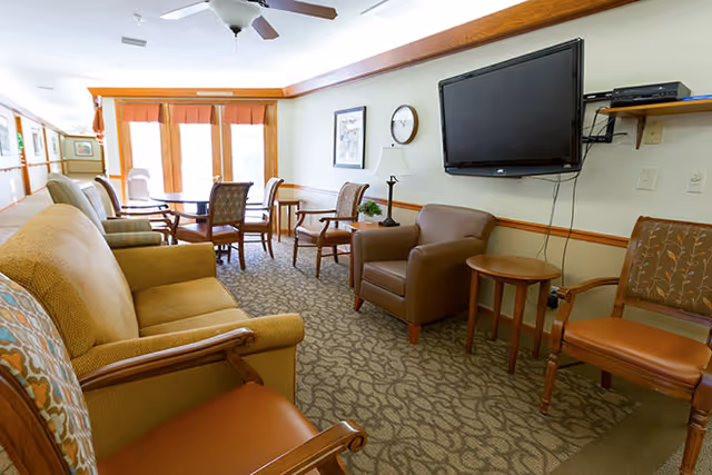 A cozy living room area in an assisted living facility with a beige sofa, several armchairs, a wall-mounted flat-screen TV, a round wooden table, and a dining table with chairs near large windows with orange curtains letting in natural light.