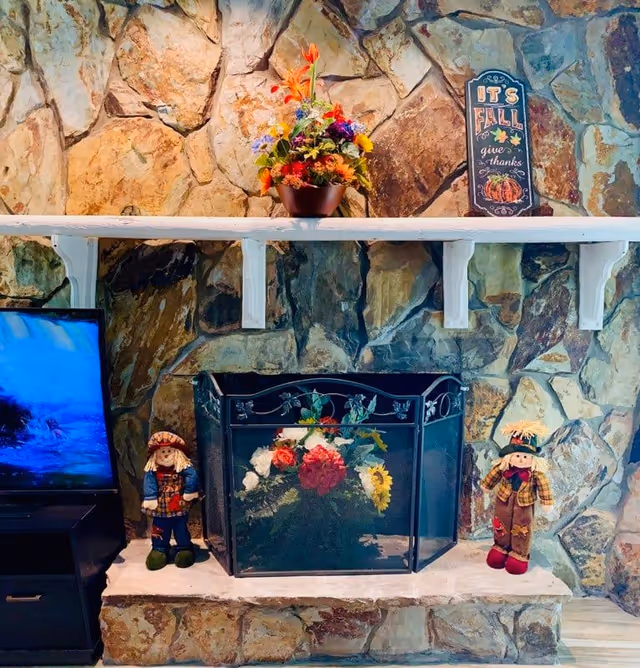 Stone fireplace with a black metal screen decorated with a floral arrangement behind it. On the mantel above, there is a colorful flower pot and a decorative sign that reads 'It's Fall give thanks' with autumn-themed illustrations. On either side of the fireplace are two small scarecrow dolls, one on the left and one on the right. To the left of the fireplace is a TV on a black stand.