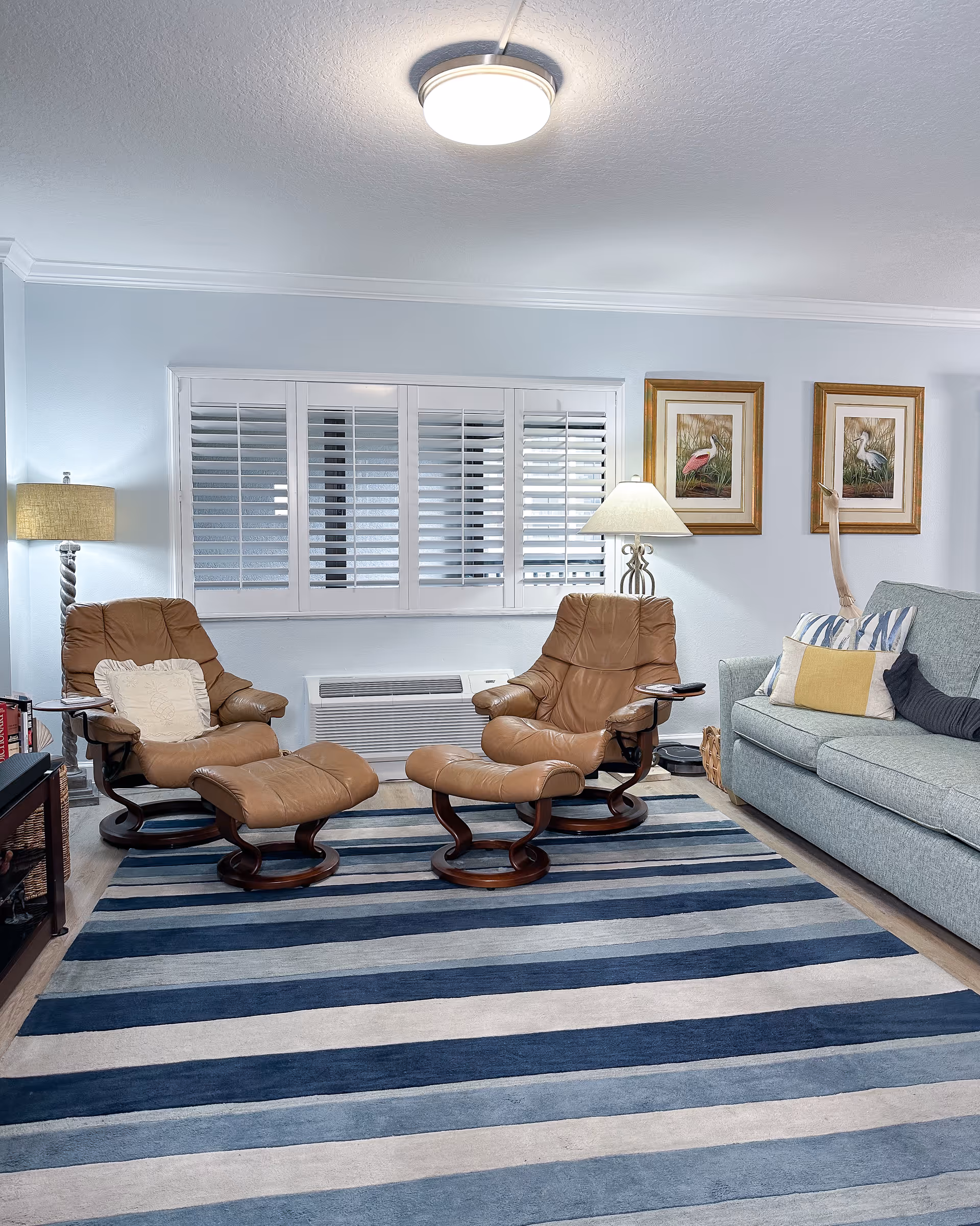 Two tan leather recliners with matching ottomans face a striped blue rug in a living room with a sofa, lamps, window shutters and framed bird prints.