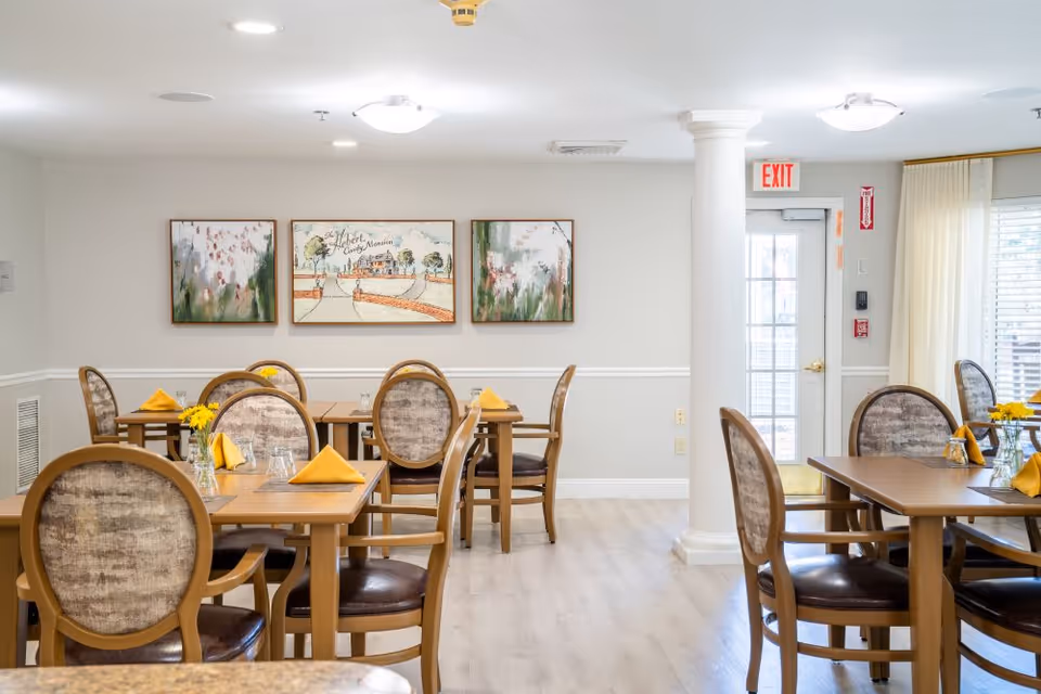 Well-lit senior dining room with wooden tables and upholstered chairs set with yellow napkins and small flower vases.
