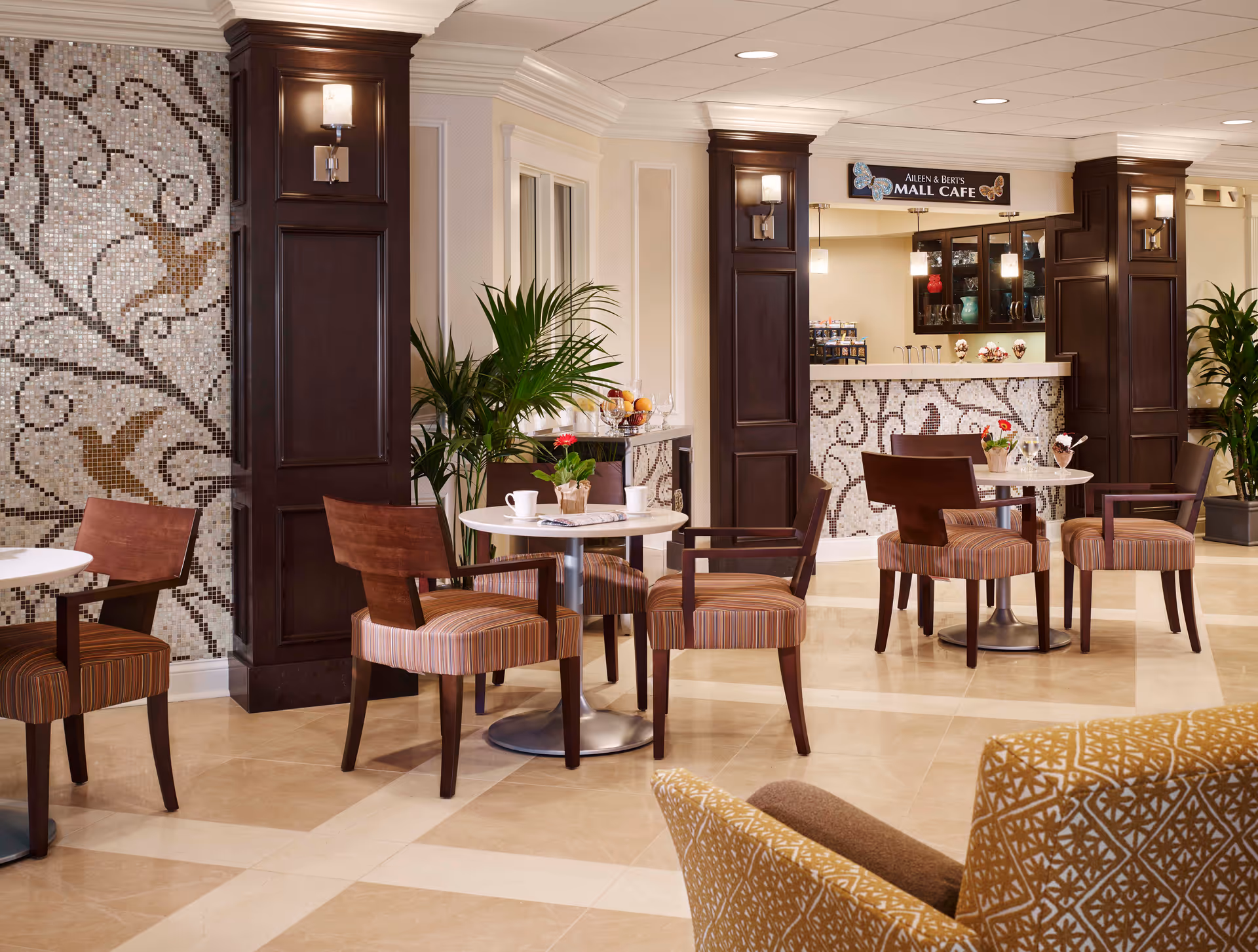 Bright, upscale indoor cafe area with round tables, striped armchairs, mosaic-tiled walls and a service counter.