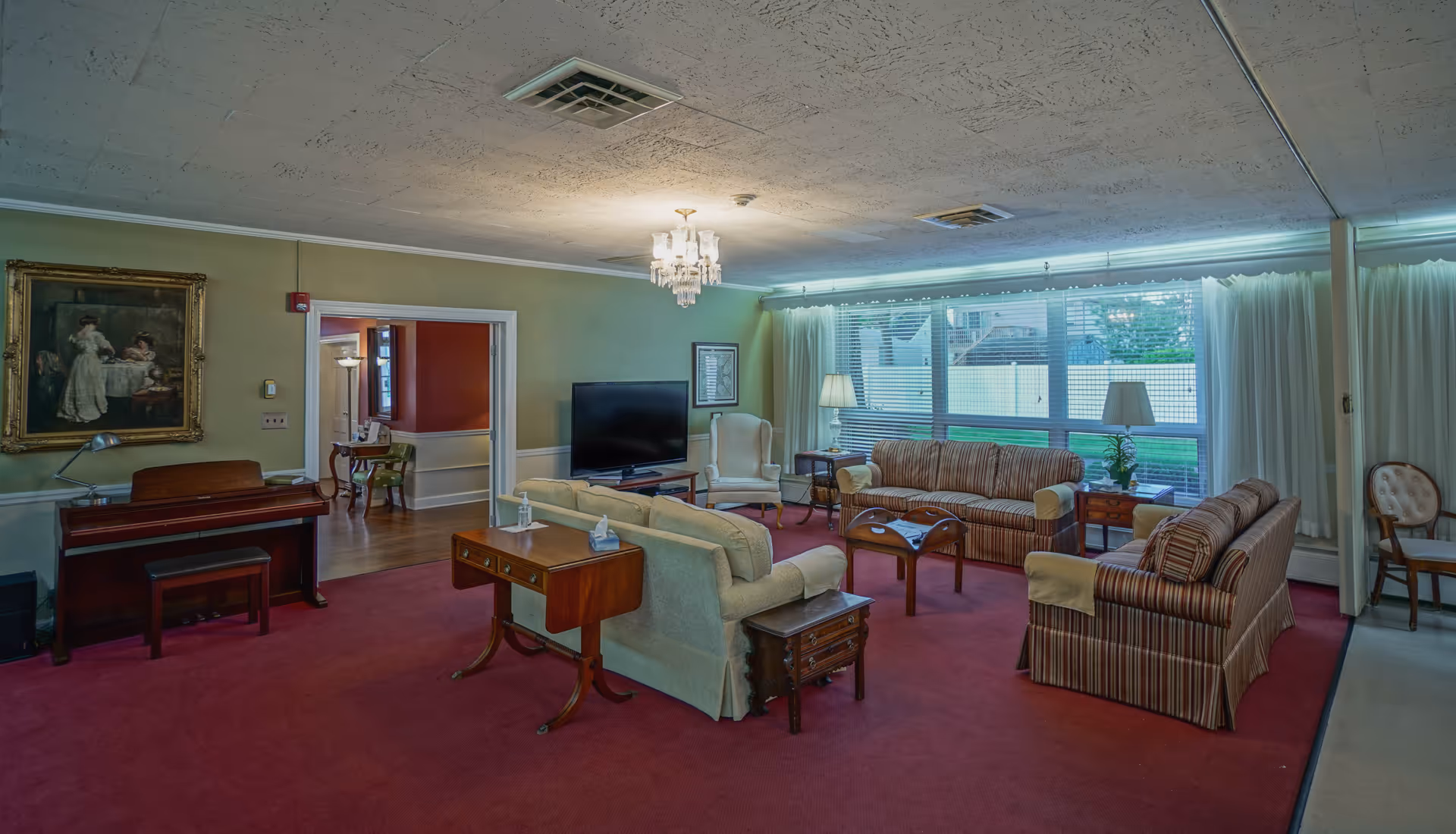 A spacious living room with red carpet, multiple sofas and armchairs arranged around a wooden coffee table. There is a piano against one wall with a painting above it, a flat-screen TV on a stand, and large windows with white curtains letting in natural light. The room has a chandelier and several lamps providing additional lighting.