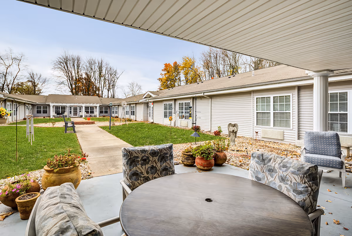 Outdoor patio area at Bennett Place with a round table and cushioned chairs under a covered roof. The patio overlooks a courtyard with a concrete walkway, green grass, potted plants, and a building with multiple windows and doors. Trees with autumn foliage are visible in the background.