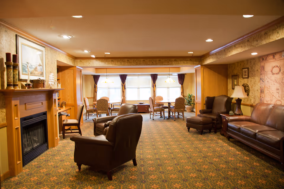 A cozy senior living common area with leather armchairs and a sofa arranged around a wooden fireplace. The room has patterned carpet, wallpapered walls, and a large window with curtains letting in natural light. There are tables and chairs near the window for dining or activities, and decorative items including a lamp, framed pictures, and candles on the mantel.