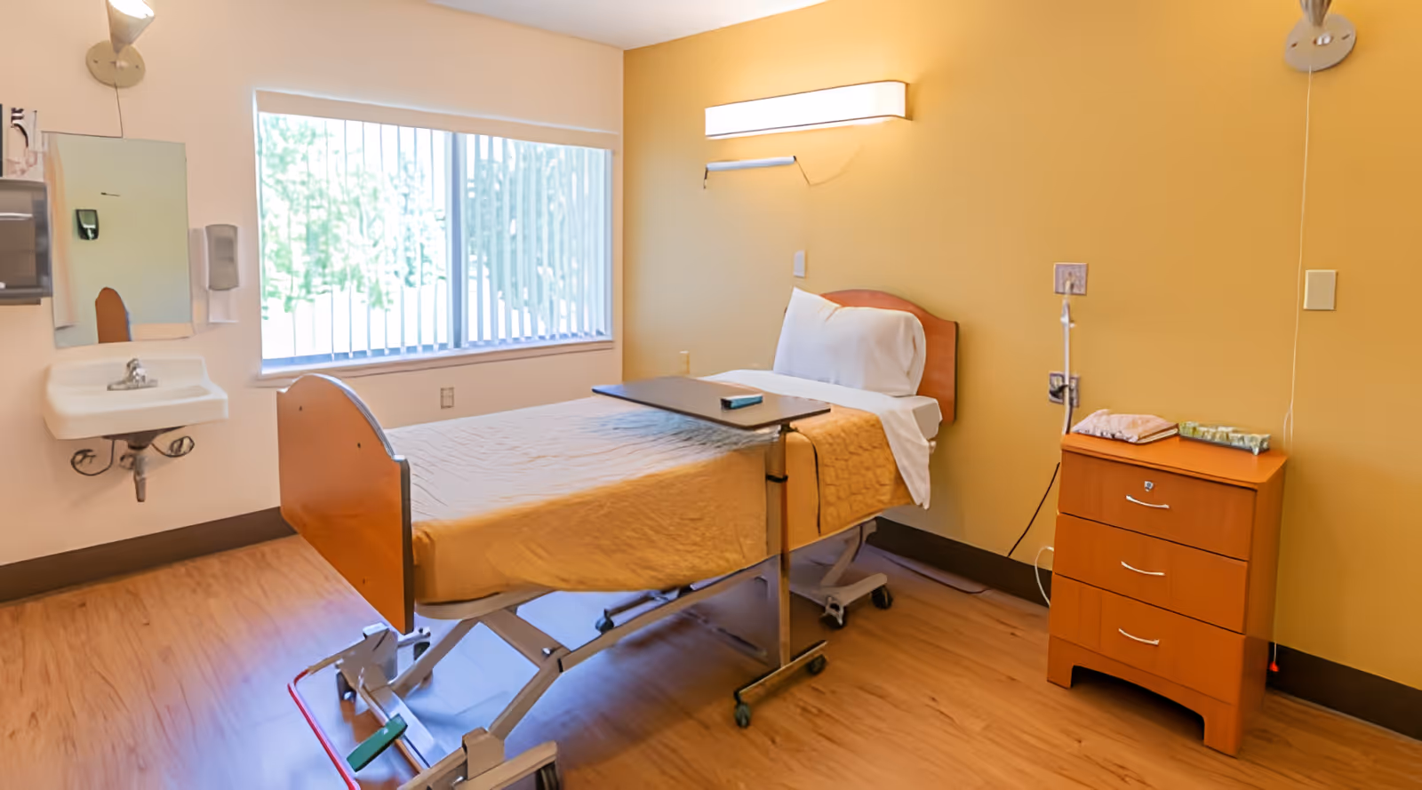 Single-occupancy nursing room with an adjustable bed, overbed table, bedside cabinet, sink, and large window.