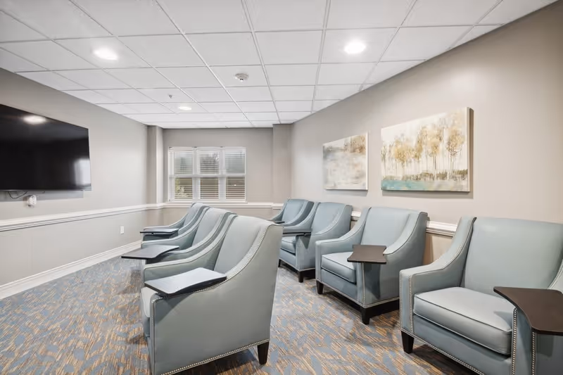 A small lounge area with six light blue armchairs arranged in two rows facing a wall-mounted flat screen TV. Each chair has a small attached black tray table. The room has beige walls with white trim, a patterned carpet, two landscape paintings on the wall, and a window with blinds letting in natural light.