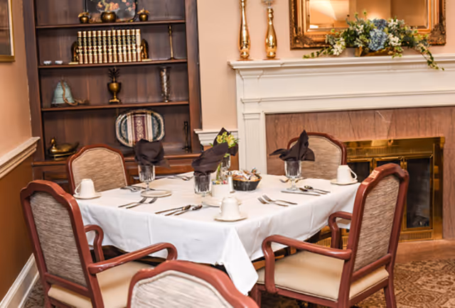 A dining room table set for six with white tablecloth, folded black napkins in glasses, cups, and silverware. The room features a fireplace with a floral arrangement on the mantel and a wooden bookshelf filled with books and decorative items in the background.