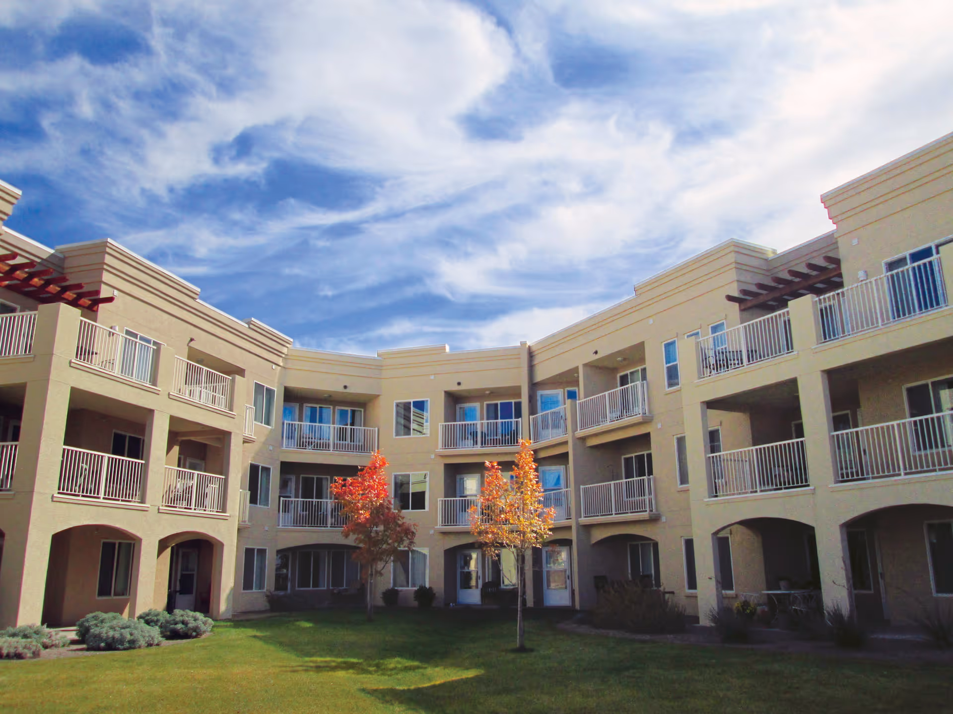 Exterior view of a three-story residential building with balconies overlooking a grassy courtyard with a few small trees under a partly cloudy sky.