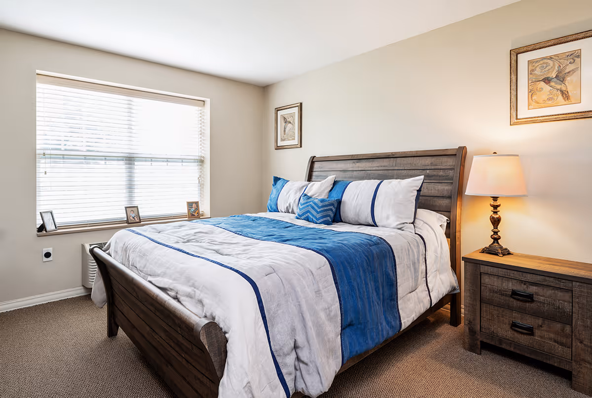 A cozy bedroom with a wooden bed frame and a neatly made bed featuring white and blue bedding. There is a wooden nightstand with a lamp on the right side of the bed. The room has beige walls with two framed bird pictures hanging above the bed. A large window with white blinds lets in natural light, and three small framed photos are placed on the windowsill.