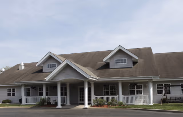 Exterior front view of a single-story building with a gray roof and white siding. The building features a covered entrance supported by white columns, multiple windows, and a small porch area with railings. There is a paved driveway or parking area in front and some landscaping with bushes and grass.