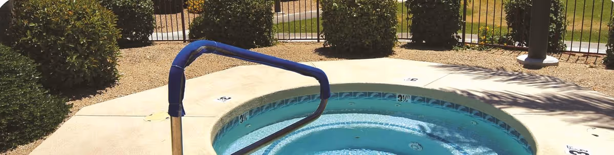 Outdoor hot tub or spa with metal handrail surrounded by concrete decking, bushes, and a black metal fence with a grassy area and trees in the background.