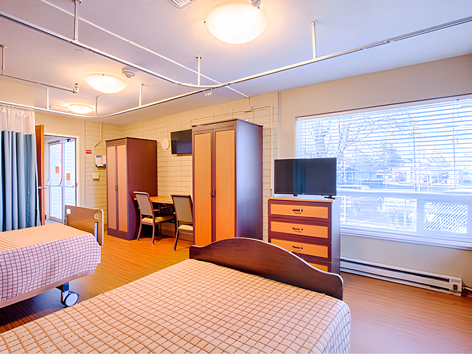 A bright room with two single beds covered in checkered bedspreads, a wooden dresser with a TV on top, two wardrobes, a small table with two chairs, and large windows with blinds letting in natural light.