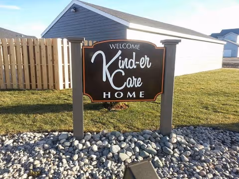 A freestanding sign reading "Welcome Kind-er Care Home" sits on rocks and grass in front of residential buildings.