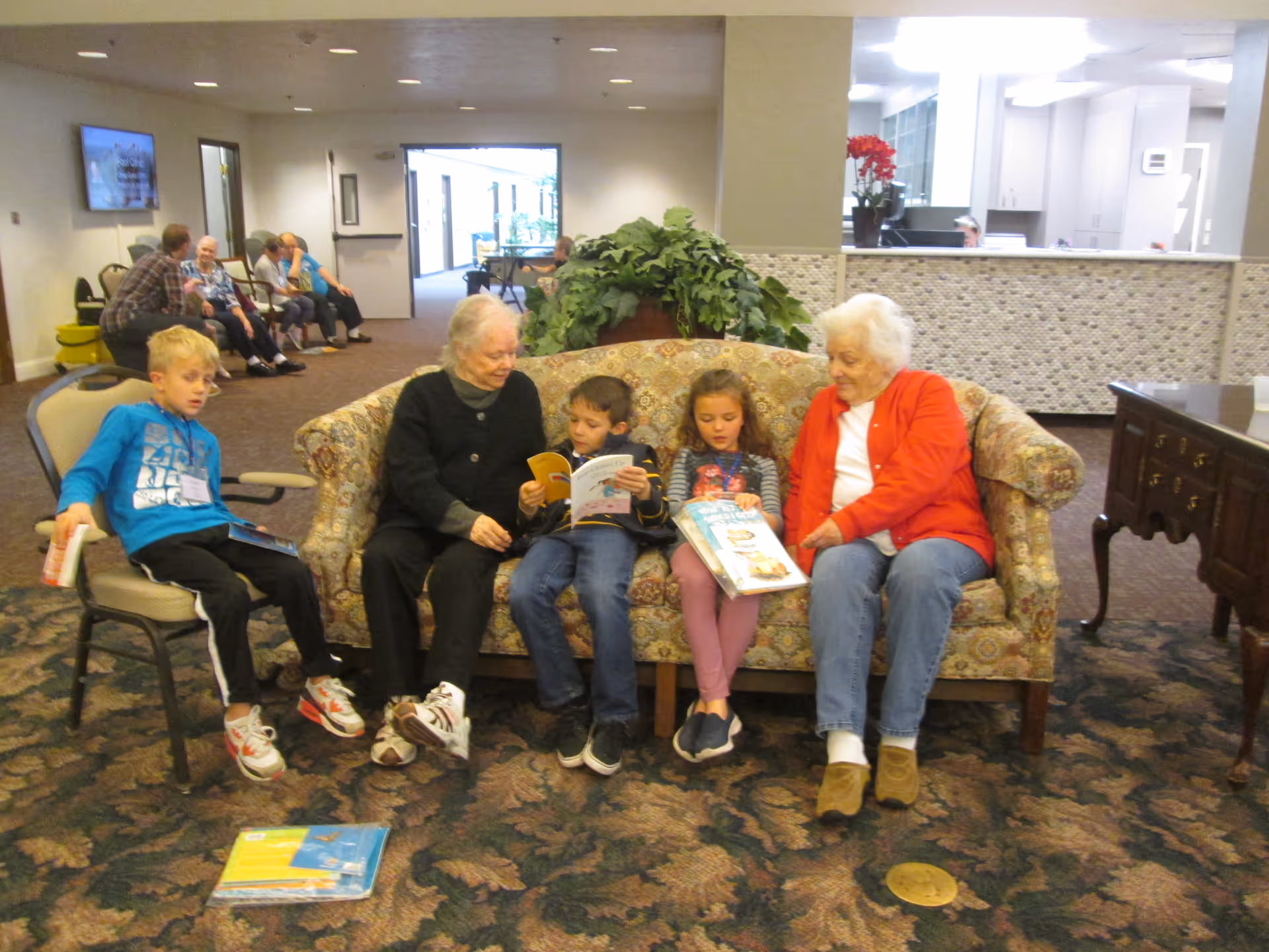 Two elderly women and two children sitting on a patterned sofa in a common area of an assisted living facility, reading books together. Another child sits on a chair nearby holding a book. In the background, several elderly people are seated along the wall, and a reception area is visible.