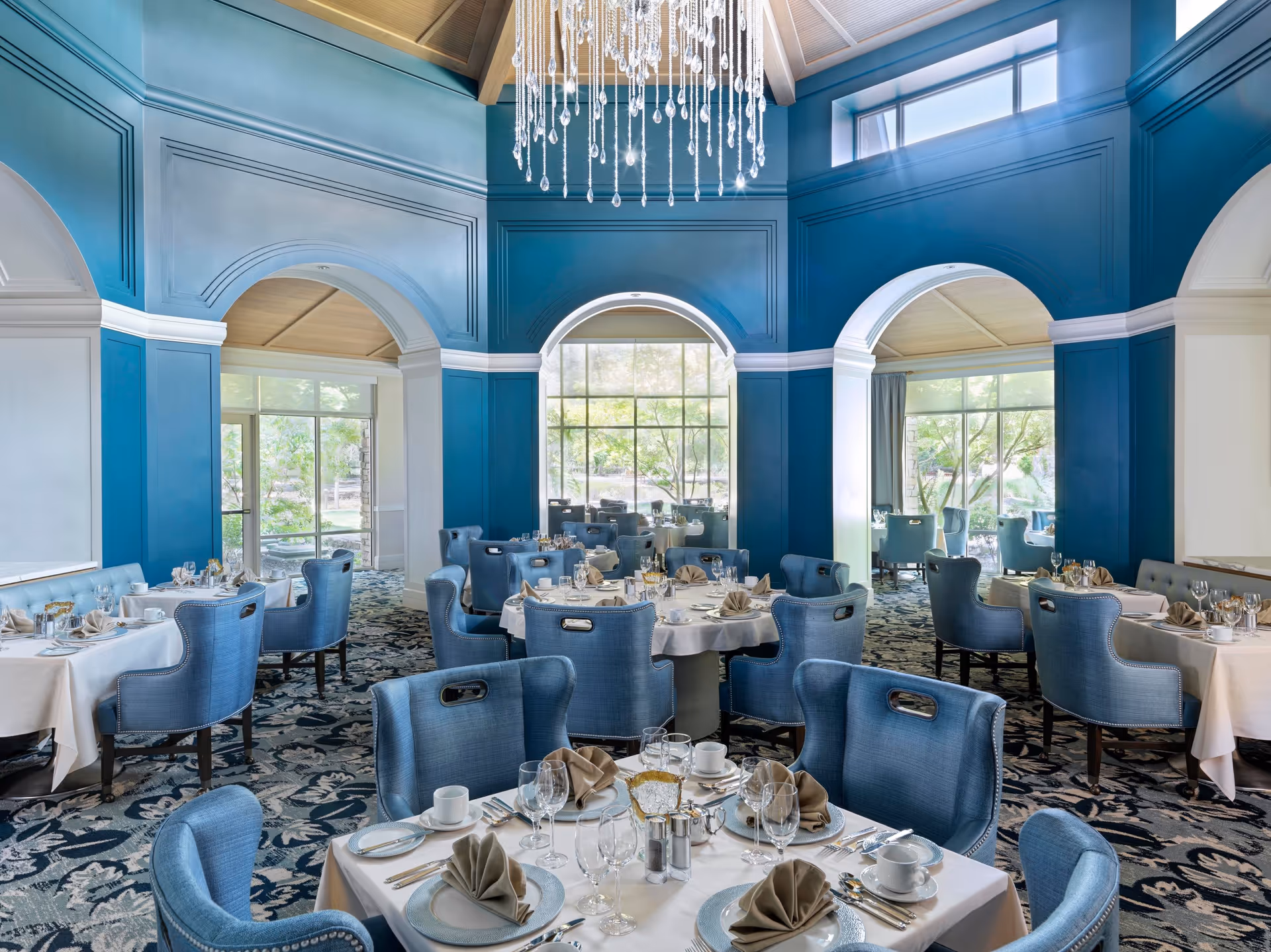 Elegant dining room with blue upholstered chairs and white tablecloths on round and rectangular tables. The room features large arched windows, blue walls with white trim, a patterned carpet, and a modern chandelier hanging from a high ceiling.
