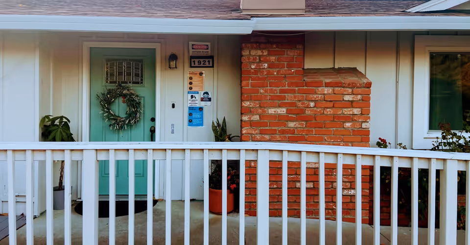 Mint green front door with wreath, brick wall, porch railing, and potted plants.