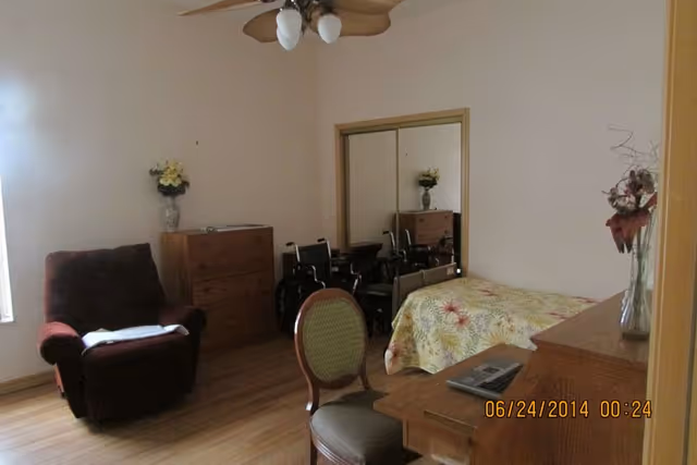 A bedroom in an assisted living facility featuring a bed with a floral bedspread, a wooden desk with a laptop, a wooden dresser, a green cushioned chair, a maroon recliner chair, and a wheelchair near a mirrored closet. The room has light-colored walls and wooden flooring.