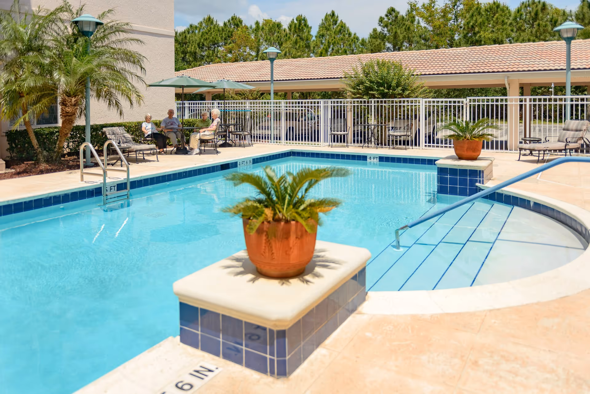 Outdoor swimming pool area with clear blue water, surrounded by a tiled deck. There are potted plants on tiled pillars near the pool steps. In the background, four elderly people are sitting and chatting under green patio umbrellas with chairs and tables. The area is fenced and has trees and shrubs around it.