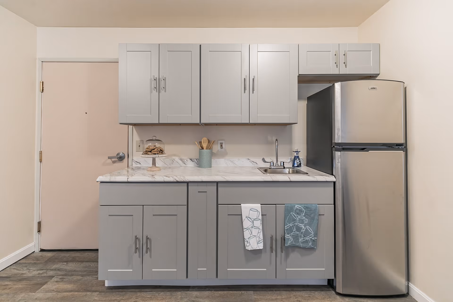 A small kitchen area with light gray cabinets, a marble countertop, a stainless steel refrigerator, a sink with a soap dispenser, and two dish towels hanging on the cabinet doors. There is a door to the left of the kitchen setup and wooden flooring.