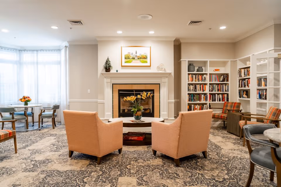 A cozy senior living common area featuring two peach-colored armchairs facing a fireplace with a floral arrangement on the coffee table in front. The room has a patterned carpet, bookshelves filled with books, and additional seating including plaid and gray chairs. Large windows with sheer curtains allow natural light to fill the space.