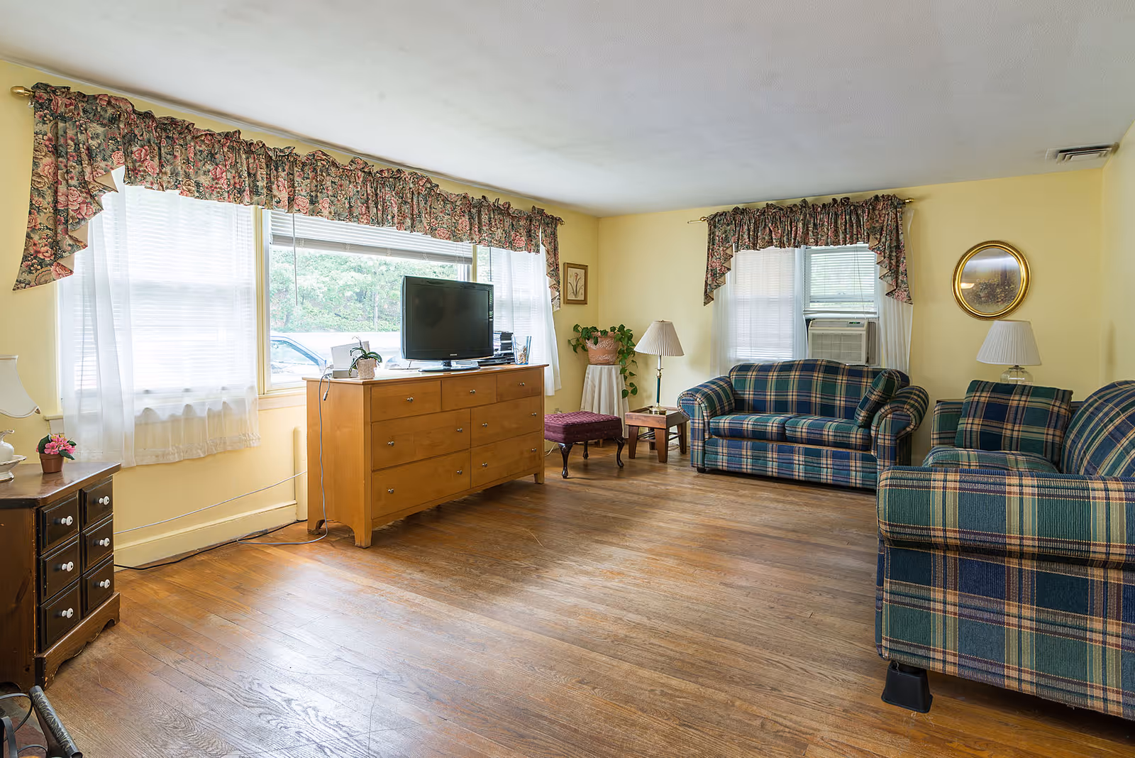 Bright living room with plaid sofas, a wooden dresser with a TV, hardwood floors and floral valance curtains.