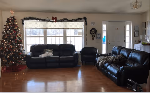 A living room with two black leather sofas and one black armchair arranged around a large window decorated with garland and a red bow. A decorated Christmas tree with lights and ornaments stands to the left. The room has wooden flooring and a white front door with glass panels on the right side. There are some framed pictures and decorations on the walls.