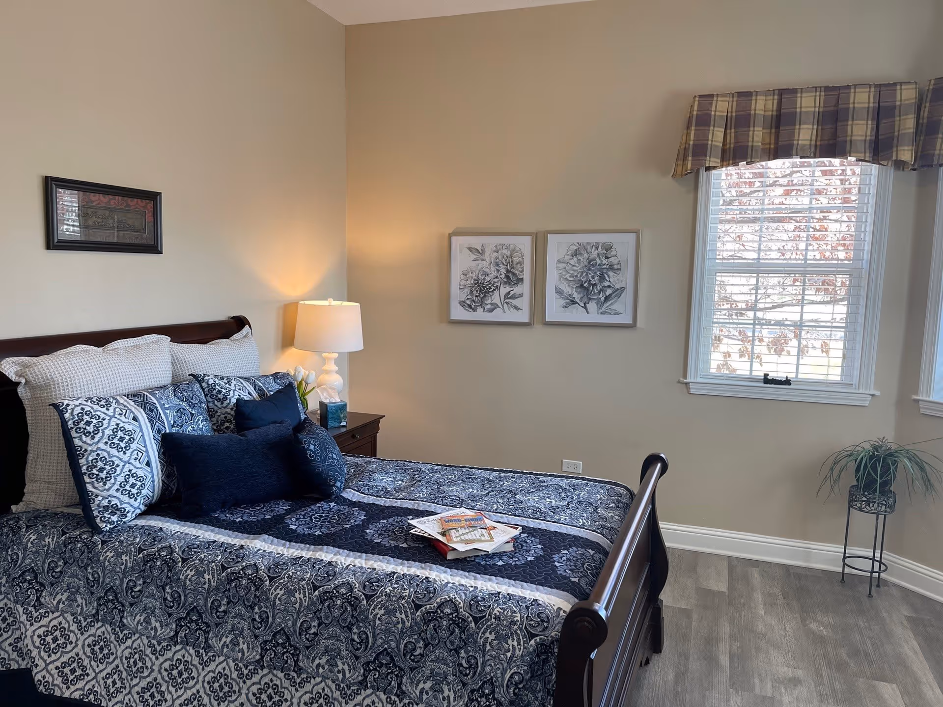 Well-decorated bedroom with a patterned blue bedspread, pillows, a nightstand with lamp, wall art, and a window with a plaid valance.