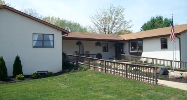 Single-story building exterior with a ramped entrance, porch seating, and an American flag on a grassy lawn.