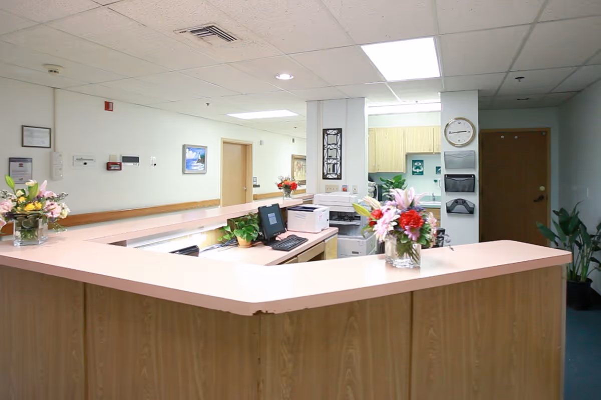 A reception/nursing station with a curved countertop, computer, printer, floral arrangements, and a hallway and doors in the background.