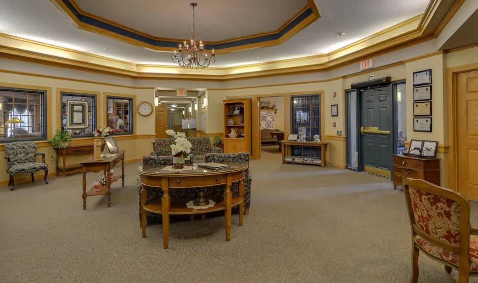 A spacious and warmly decorated common area in a senior living facility with floral upholstered chairs, wooden tables, a chandelier, and framed certificates on the wall near a green exit door.