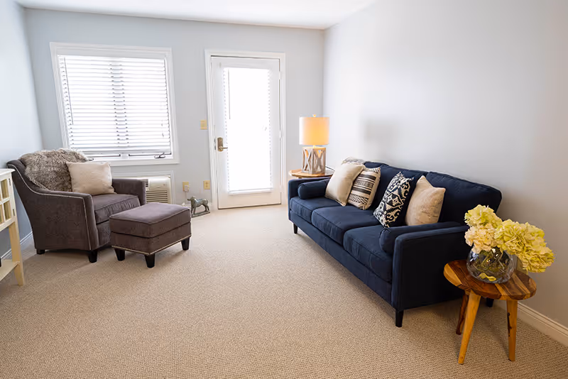 A bright living room with a navy blue sofa adorned with multiple decorative pillows, a wooden side table with a vase of yellow flowers, a round wooden lamp table with a lamp, and a gray armchair with a matching ottoman near a window and a glass door with blinds.