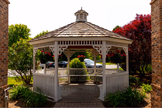 A white wooden gazebo with a shingled roof situated outdoors between two brick pillars. The gazebo is surrounded by green bushes, plants, and trees with red and green foliage. Several cars are parked in the background on a paved area.