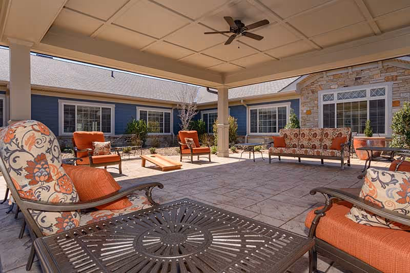 Covered outdoor patio area with cushioned chairs and sofas featuring floral and orange patterns, a ceiling fan, and a view of a courtyard with blue siding and stone exterior walls of the building.
